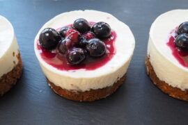 Close up of three smal round cheesecakes with a flapjack base and topped with blueberries and blueberry sauce on a dark slate background