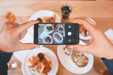 someones hands holding a phone in frojnt of a tabkle of food taking a picture of it to capture the future of food