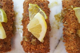 Close up of a rectangular flapjack drizzkled with gin and tonic flavoured syrup and topped with a quarter slice of lemon and lime. On a white marbled background