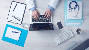 doctors hands usng a laptop on a desk with other medical admin files