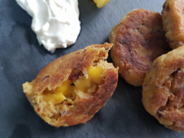 Close up of 4 vegan cheese falafels on a grey slate board with lemon slices and a dollopof aioli inthe corner. One falafel has been halved revealing a melted vegan cheese centre