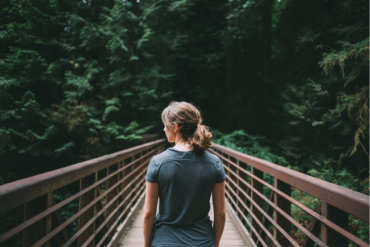 Female pictured from the waist up stood at the start of a bridge leading into a wooded area. Is is being used to suggest she is usng nature to help boost her health